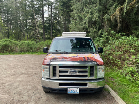 Front view of a customized Ford RV rental van with a painted hood & roof box, parked in a lush forest setting.