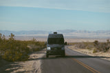 Dark Mercedes Sprinter van (RV rental) on a desert road, surrounded by bushes and distant mountains under a blue sky.