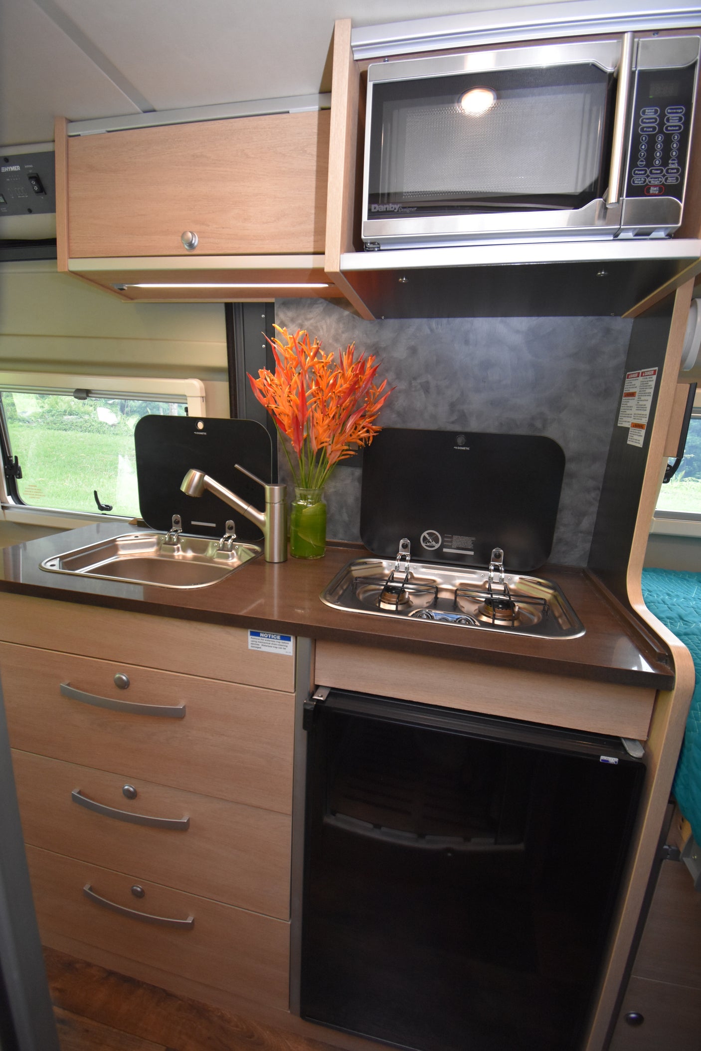 An RV rental kitchen features a sink, stove, microwave, fridge, cabinets, and a vase of orange flowers.