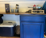 RV rental kitchenette features a portable camping stove, Nighthawk Black wine, and cooler, ideal for road trip cooking.