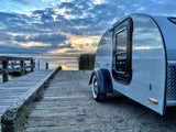 Sleek teardrop RV rental parked on a weathered wooden pier at sunrise over a calm lake.