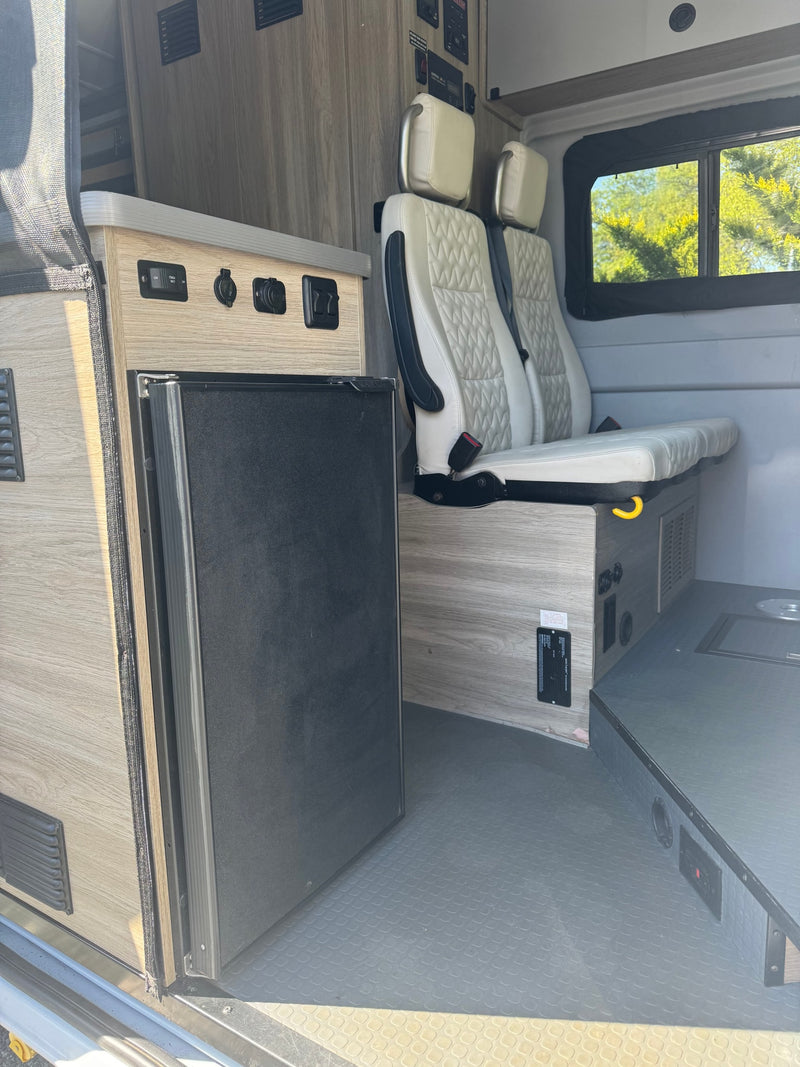 RV rental interior: light wood-look cabinets, dark fridge, power panel, and quilted passenger seats. — photo 10