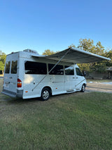 White Dodge Sprinter camper van with extended awning on grass, ready for an RV rental trip. Blue sky & trees in background.