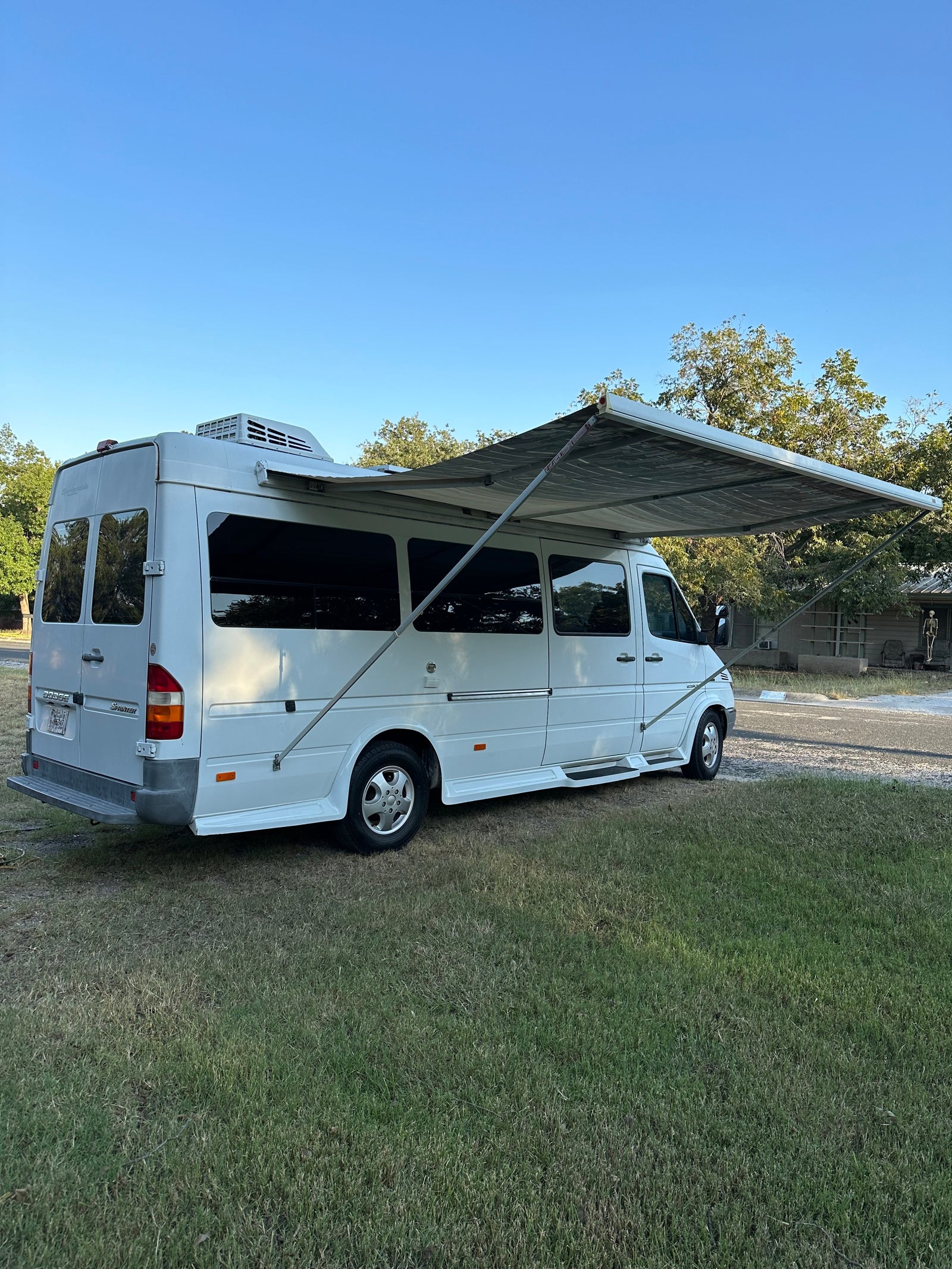 White Dodge Sprinter camper van with extended awning on grass, ready for an RV rental trip. Blue sky & trees in background.