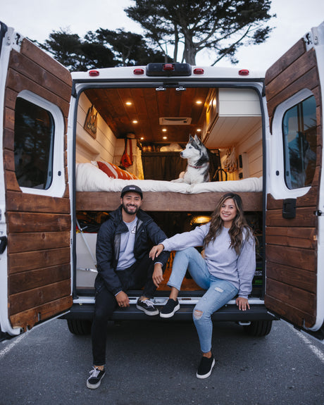 A smiling couple sits at the open back of their RV rental, with their husky relaxing on the bed inside the custom van.