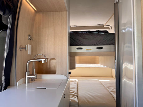 Modern RV rental interior with a compact kitchen sink, wood paneling, and a bright, cozy sleeping area, including an upper bunk.