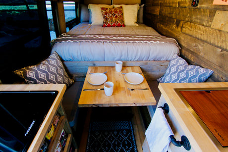 Cozy RV rental interior with a bed, dining table for two, Dometic stovetop, and Ruvati sink.