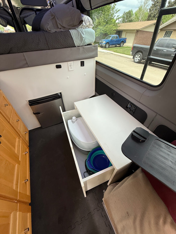 This RV rental camper van showcases a compact interior with an elevated bed, small fridge, pull-out toilet, and cabinets.