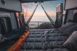 Cozy RV rental camper van bed facing an ocean sunset through screen-covered open doors, perfect for scenic retreats.