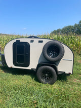 Compact grey RV rental, an off-road ready travel trailer, parked in a green field next to a cornfield under a blue sky.