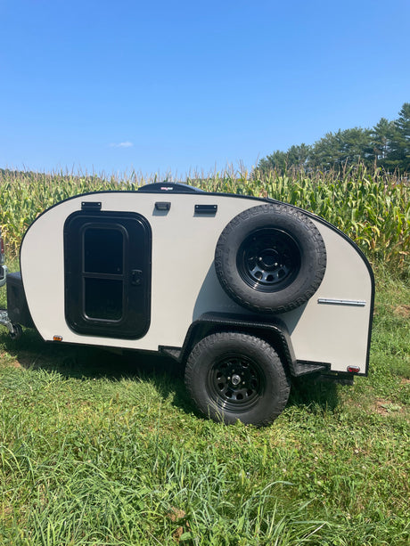 Compact grey RV rental, an off-road ready travel trailer, parked in a green field next to a cornfield under a blue sky.