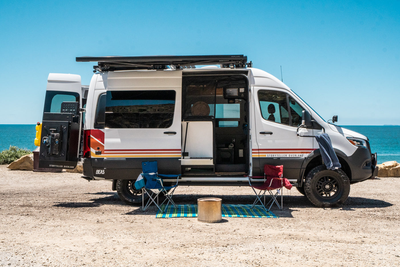 Storyteller Overland RV rental camper van parked beachside, doors open. Two chairs & fire pit set for outdoor comfort. — photo 2