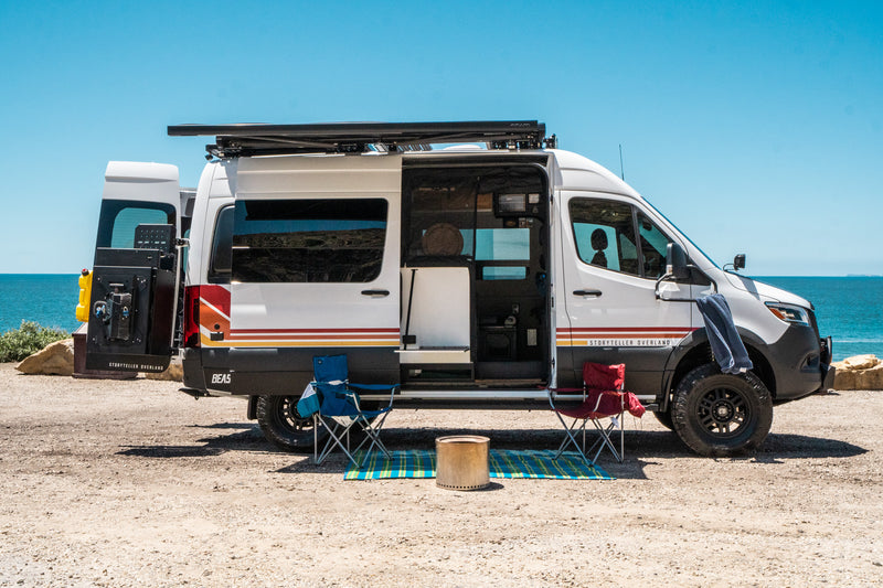 Storyteller Overland RV rental camper van parked beachside, doors open. Two chairs & fire pit set for outdoor comfort. — photo 2