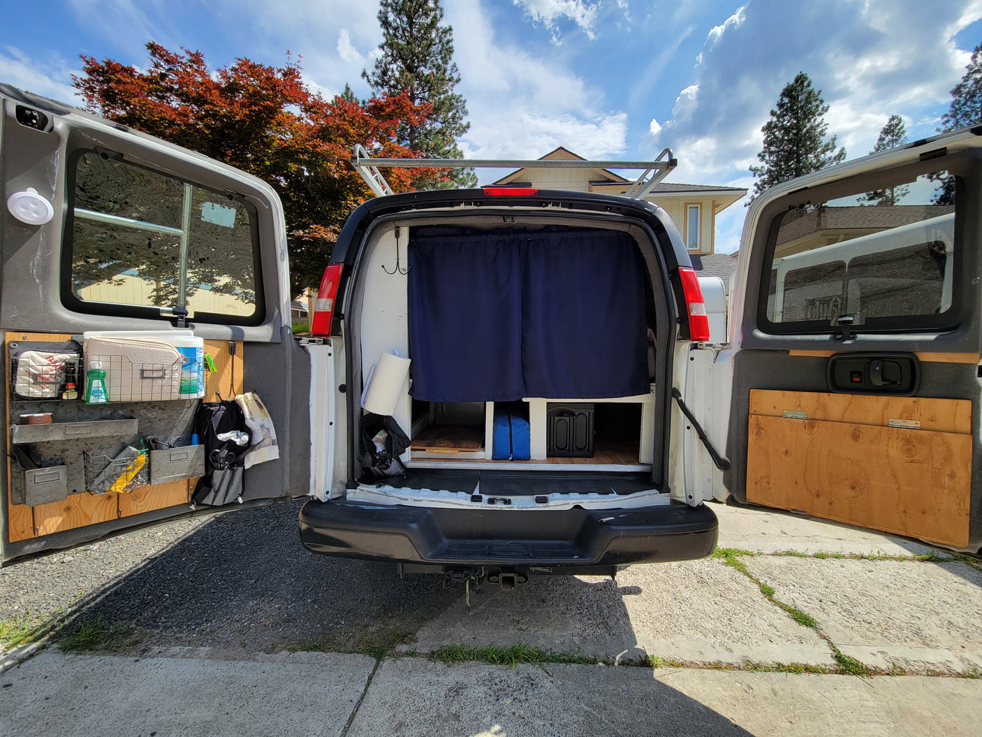 Rear view of a converted van RV rental with open doors, revealing custom storage & blue privacy curtains. Ideal for travel.