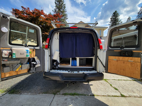 Rear view of a converted van RV rental with open doors, revealing custom storage & blue privacy curtains. Ideal for travel.