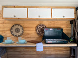 Inside an RV rental: a compact kitchen with wood paneling, white overhead cabinets, a Coleman propane stove, and dining essentials.
