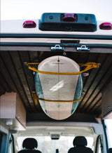 View of a surfboard strapped to the wooden ceiling inside an **RV rental** van. It's held by yellow straps, ready for travel.