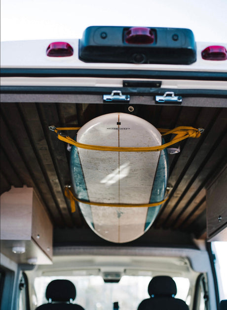 View of a surfboard strapped to the wooden ceiling inside an **RV rental** van. It's held by yellow straps, ready for travel.