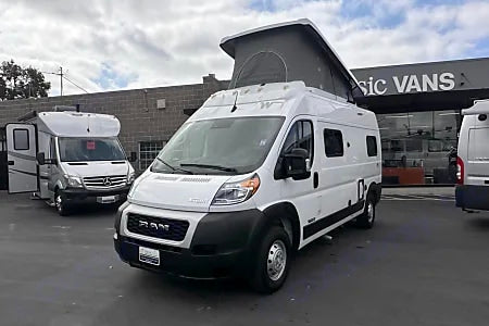Two white RVs, including a pop-top Ram ProMaster camper van, parked at an rv rental dealership.