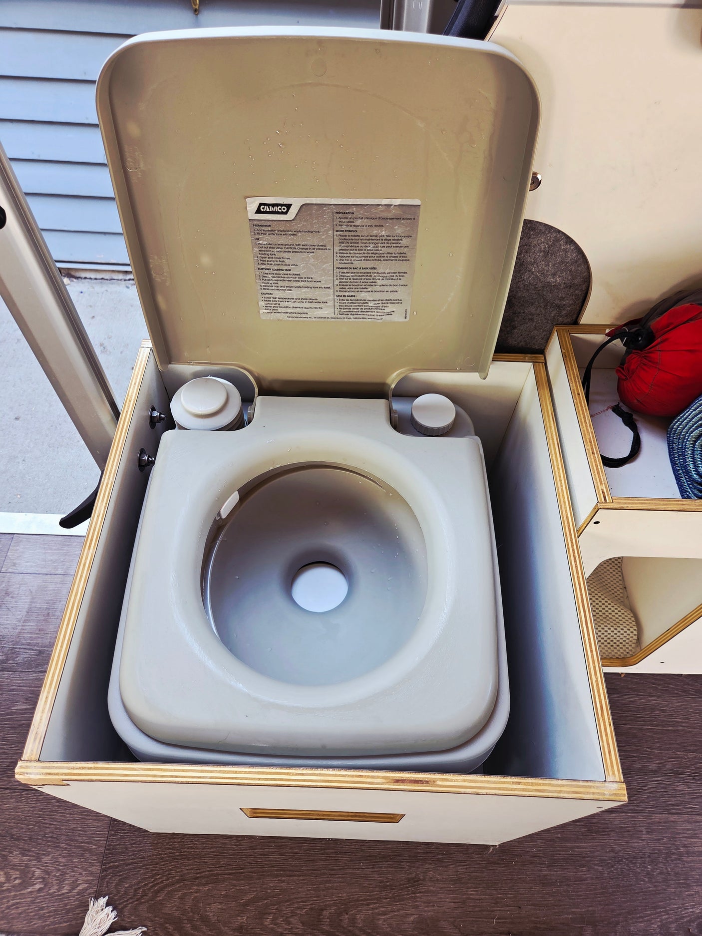 Tan portable toilet for an **rv rental** or camper van, set into a wooden box. The lid is open, showing a water-filled bowl with instructions.