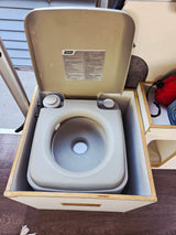 Tan portable toilet for an **rv rental** or camper van, set into a wooden box. The lid is open, showing a water-filled bowl with instructions.