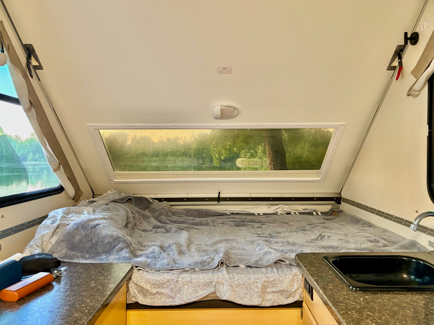Cozy RV rental interior with a comfortable bed, compact kitchen area, and a scenic window view of a lake and lush trees.