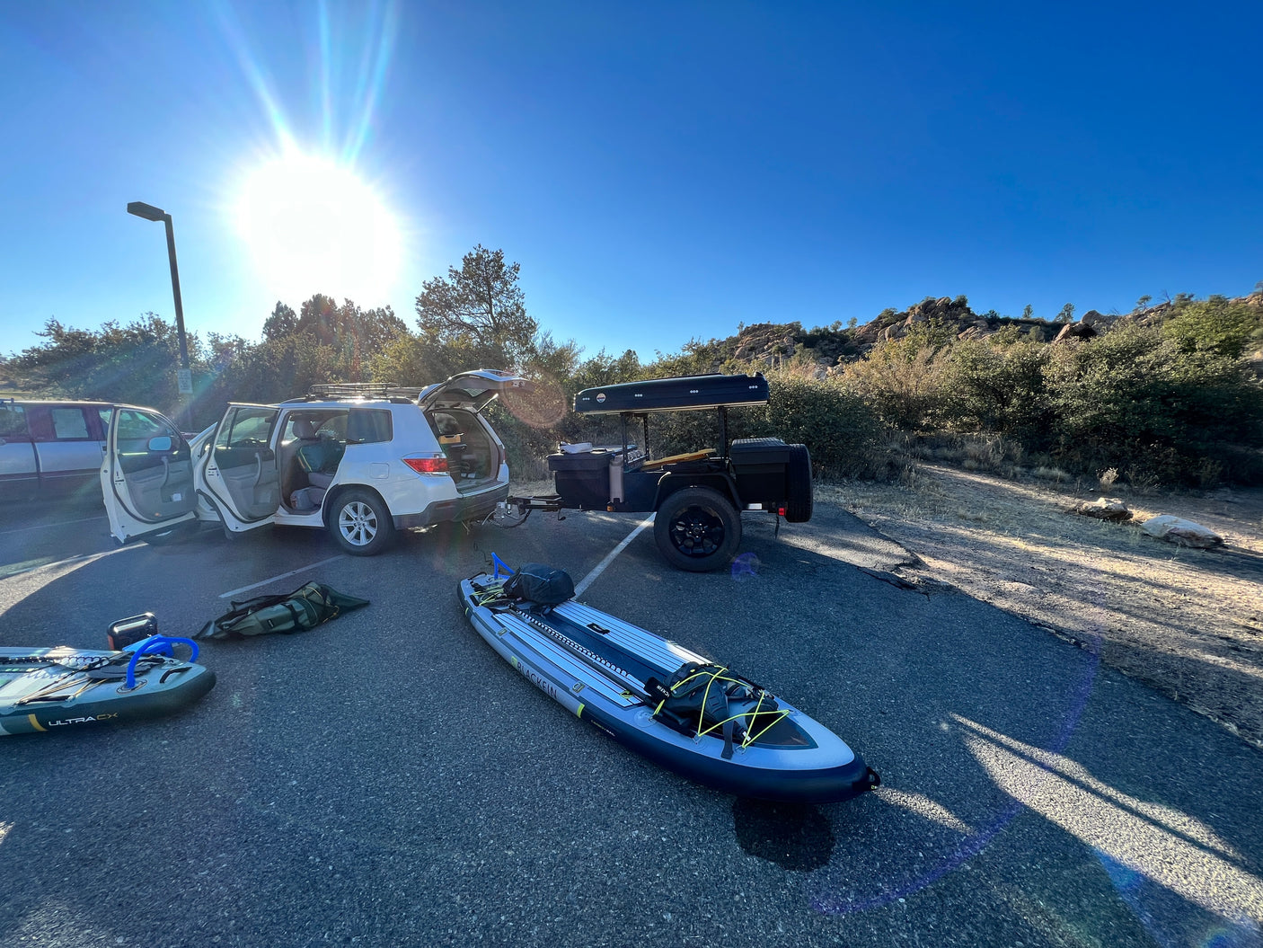 Bright sun over a parked SUV and overland trailer with rooftop tent, flanked by inflatable kayaks. Ready for outdoor rv rental trips!