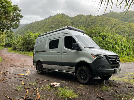 A silver Winnebago Revel **rv rental** is parked on a muddy path in lush green mountains, with chickens nearby. Perfect for adventure travel!