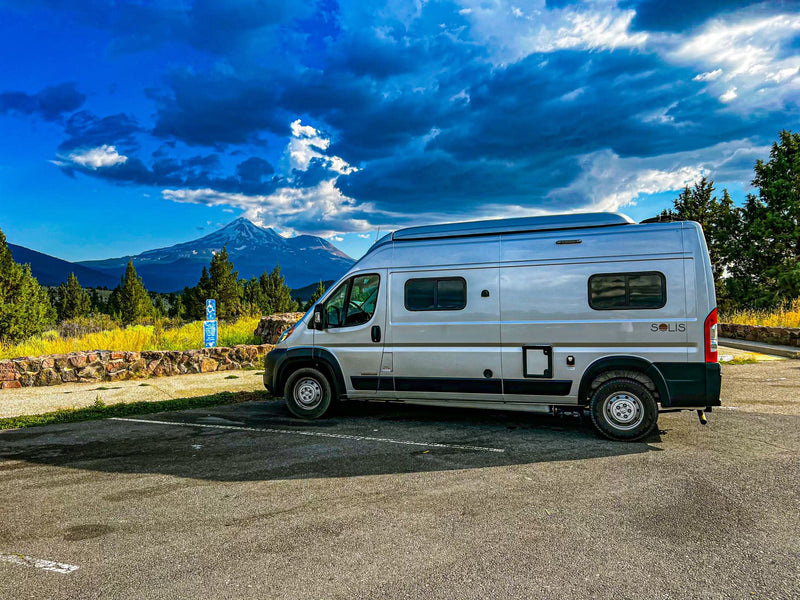 parked on a sunny dirt road.|Silver camper van RV rental parked with a majestic snow-capped mountain & dramatic sky. Perfect for scenic travels.|A silver Winnebago Solis RV rental van parked on a road — photo 2