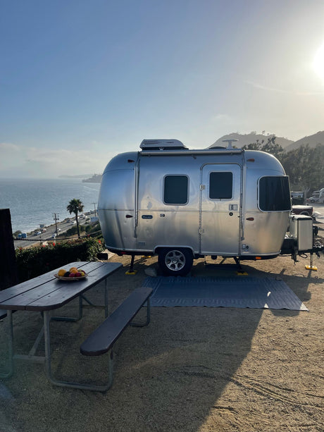 Silver Airstream RV rental at a scenic oceanfront campsite. Features picnic table, outdoor mat, and stunning coastal views.