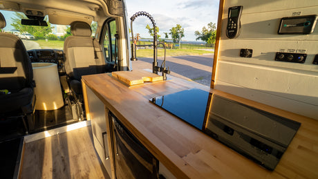 Inside an RV rental: a sleek kitchen area with wood counter, sink, induction cooktop, and control panel, looking out to nature.