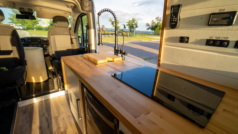 Inside an RV rental: a sleek kitchen area with wood counter, sink, induction cooktop, and control panel, looking out to nature. — photo 19