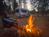 RV rental adventure: woman warms hands by a crackling campfire, her camper van parked in a forest at dusk.