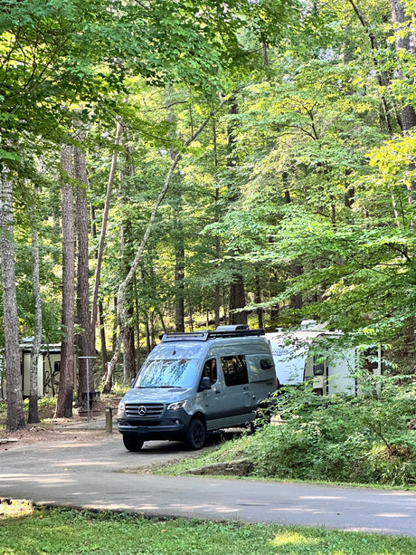 Serene campground scene: A gray Sprinter RV rental parked next to a white travel trailer amidst tall trees.