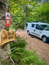 An RV rental white van is parked at Hipcamp site 11, 'Blue Jay Way.' A woodsy campsite with a fire pit and wood chips.