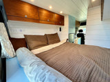 Cozy RV rental bedroom with warm wooden cabinets, white shiplap, and a comfy bed. A light blue kitchenette is visible. Ideal for travel.