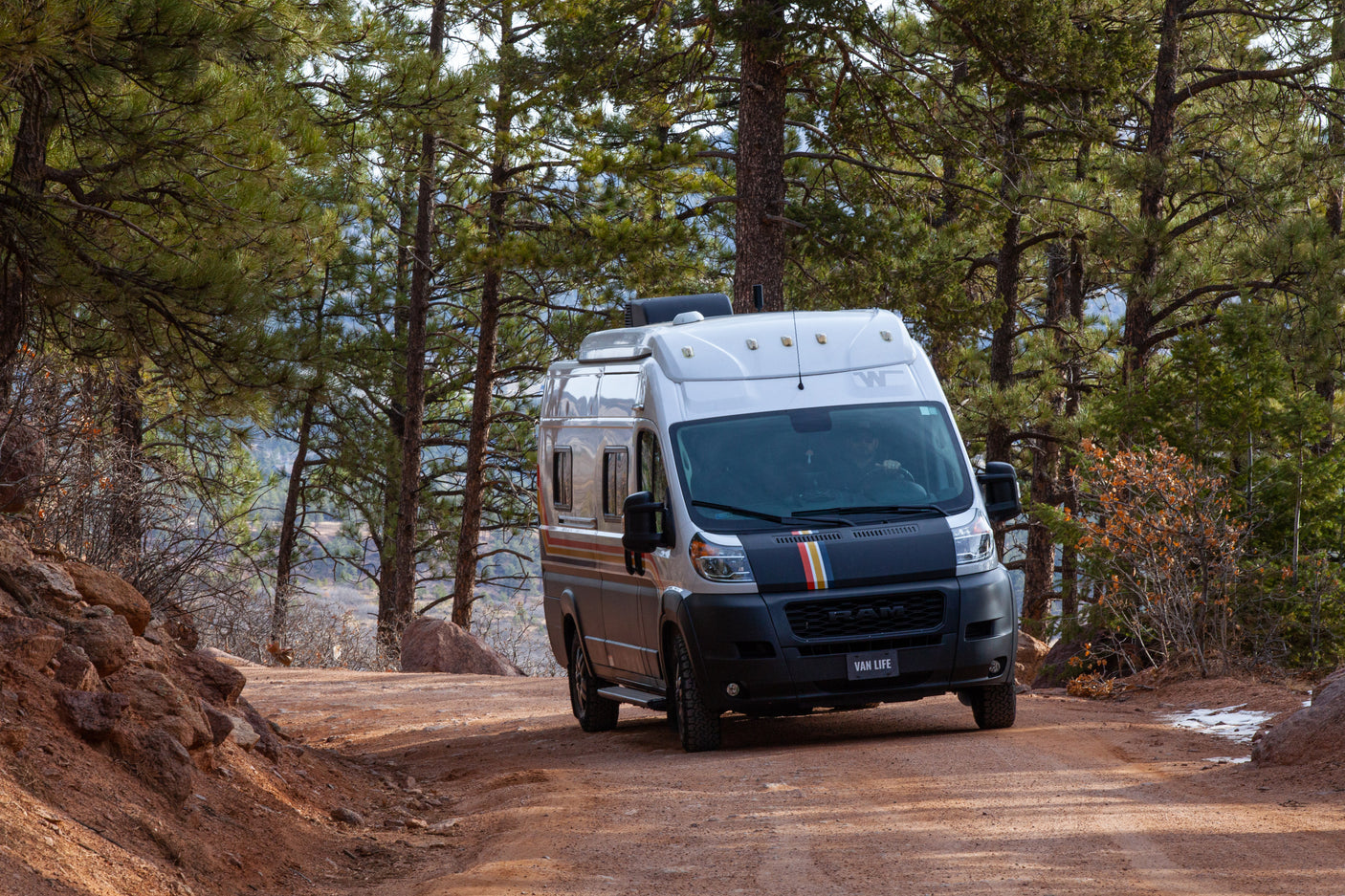 A modern camper van, ideal RV rental for off-road adventure, drives on a scenic dirt road through a pine forest.
