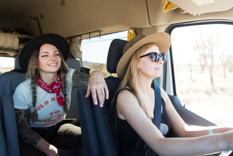 Two women enjoy an RV rental road trip; one smiles as a passenger, while the other drives through a sunny desert landscape.