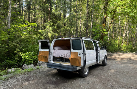 Cozy white RV rental camper van parked in a vibrant green forest, doors open revealing a bed. Perfect for a road trip!
