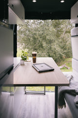 Cozy RV rental workspace: laptop, coffee, and plant on a table. Enjoy remote work with a scenic view of nature.