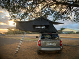 A tan Jeep Patriot with a rooftop tent (an RV rental option) set up for camping at sunset, ladder extended.