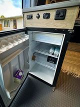 RV rental: Open compact fridge in a camper van, showing a control panel with power outlets above. Contains drinks, Half & Half, and egg holders.