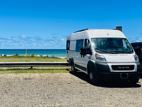 White Ram ProMaster RV rental van on gravel, facing a beautiful blue beach with ocean under a clear sky.