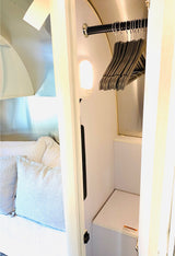 Inside a modern RV rental: a compact bedroom with light bedding, and an open wardrobe with hangers and an interior light. Perfect for travel.
