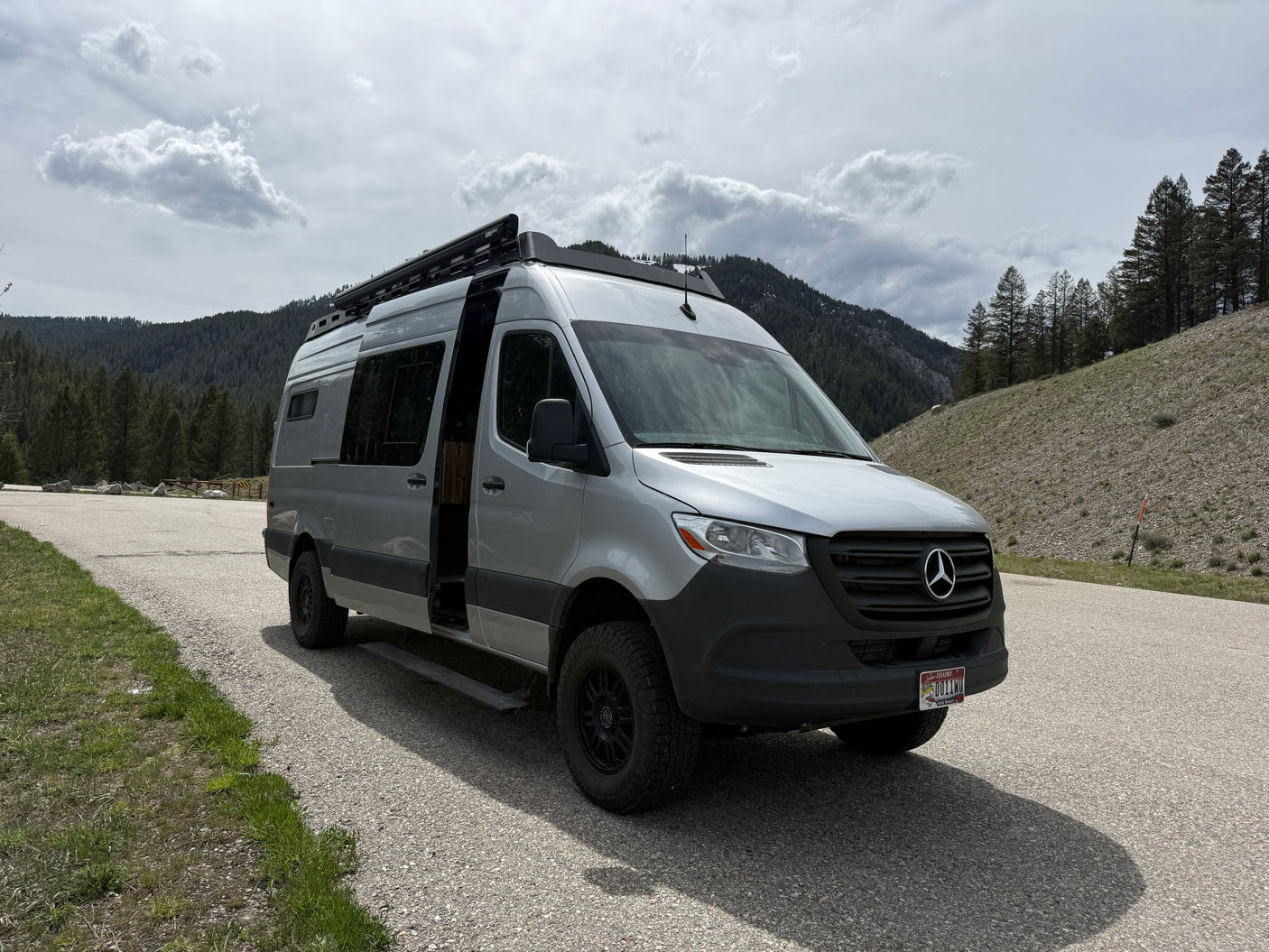 Spacious silver rv rental van (Mercedes Sprinter) with an open door parked on a mountain road amidst pines under cloudy skies.