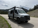 Spacious silver rv rental van (Mercedes Sprinter) with an open door parked on a mountain road amidst pines under cloudy skies.