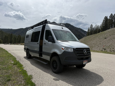 Spacious silver rv rental van (Mercedes Sprinter) with an open door parked on a mountain road amidst pines under cloudy skies.