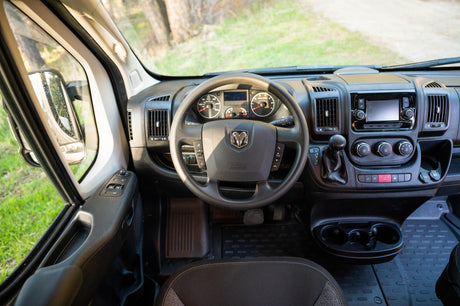 Inside an RV rental: driver's cabin with steering wheel, dashboard, controls, and a view of nature through the windshield.