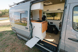 Stylish RV rental: Grey camper van interior with kitchen, flip-out table, and living area, parked by a scenic lake.