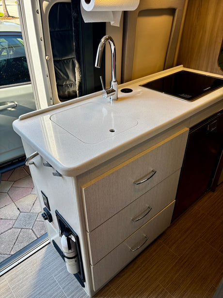 RV rental kitchen: modern sink, induction stove, built-in drawers, and a fire extinguisher mounted by the door.